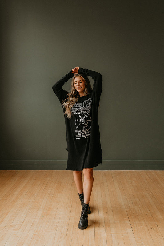 Woman wearing a black Led Zeppelin graphic dress standing in a room with a wooden floor and gray wall.