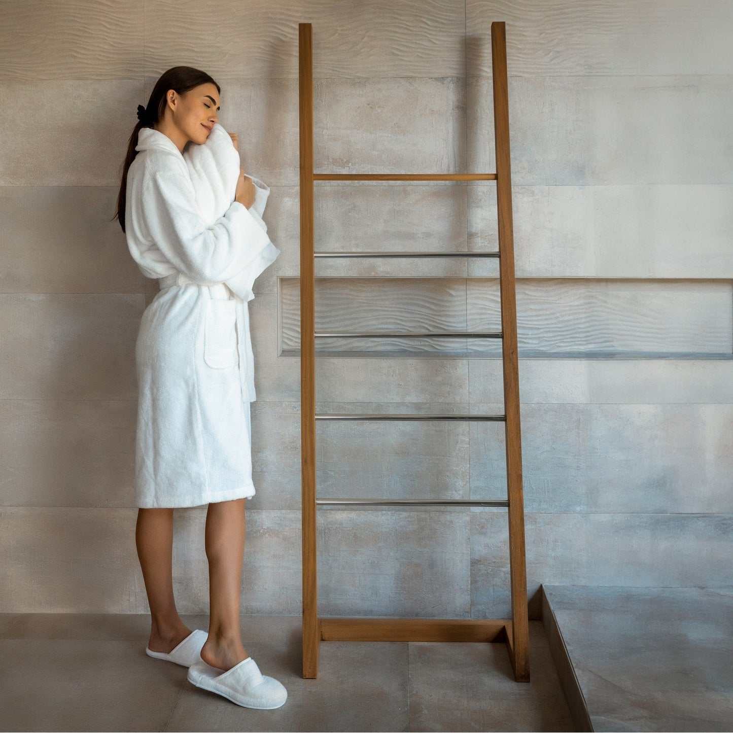 Woman in a white robe and slippers standing next to a wooden towel ladder in a bathroom.