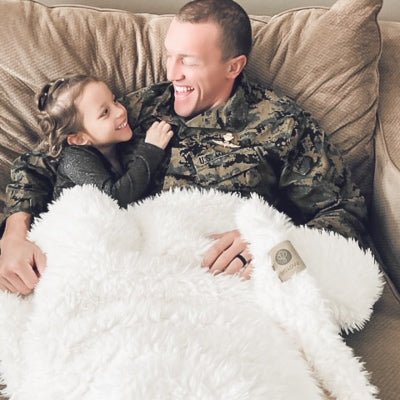 Man in military uniform sitting on a couch with a child, both lying on a fluffy white blanket.
