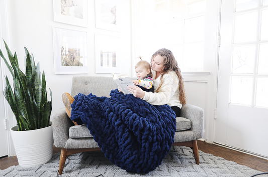Woman sitting on a gray chair holding a baby, with a large navy blue knitted blanket draped over her lap in a bright room.