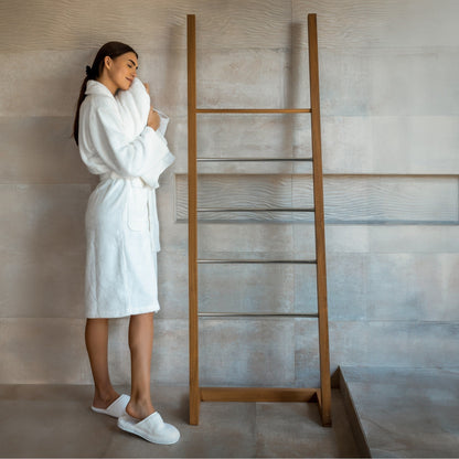 Woman in a white robe and slippers standing next to a wooden towel ladder in a bathroom.