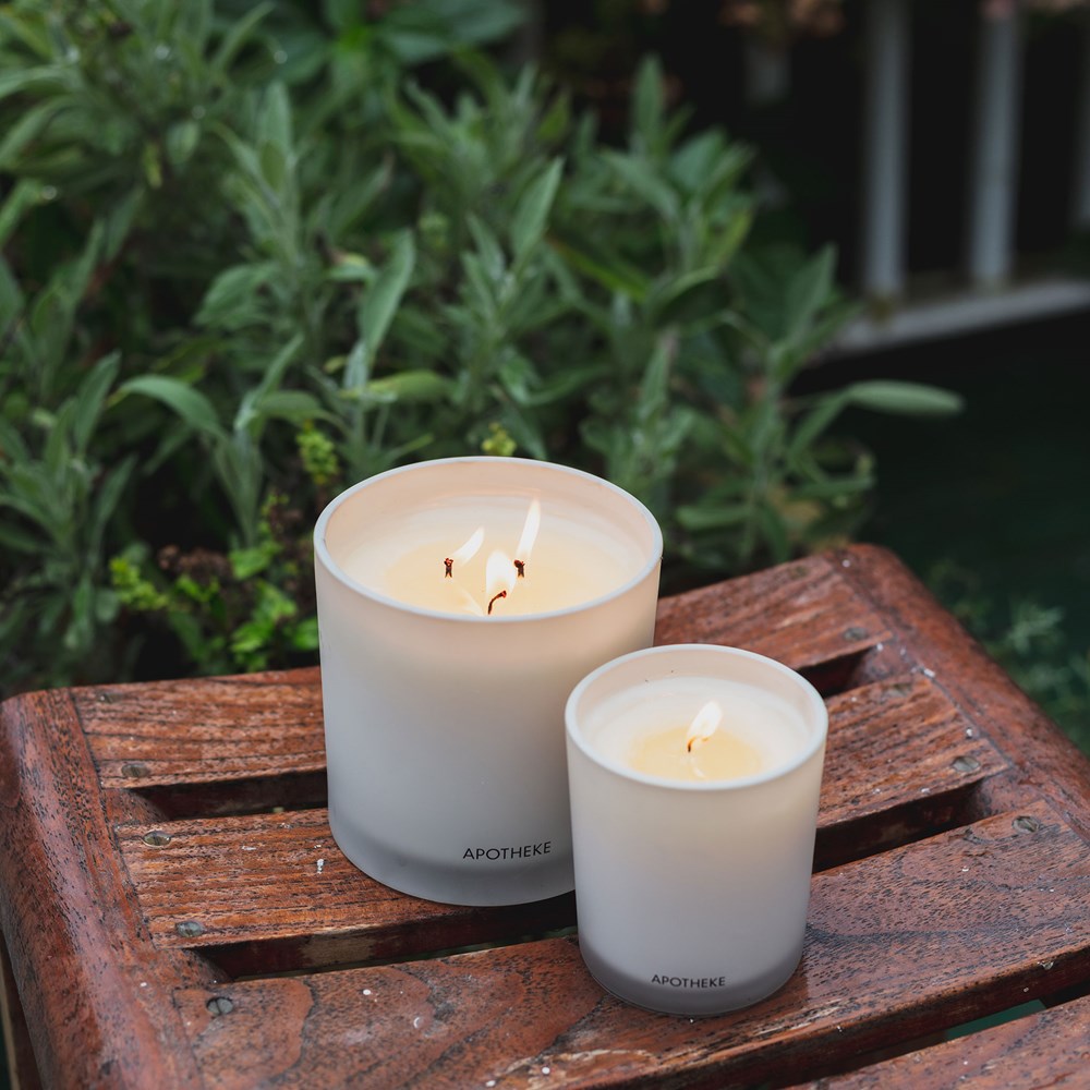 Two white candles on a wooden surface with greenery in the background