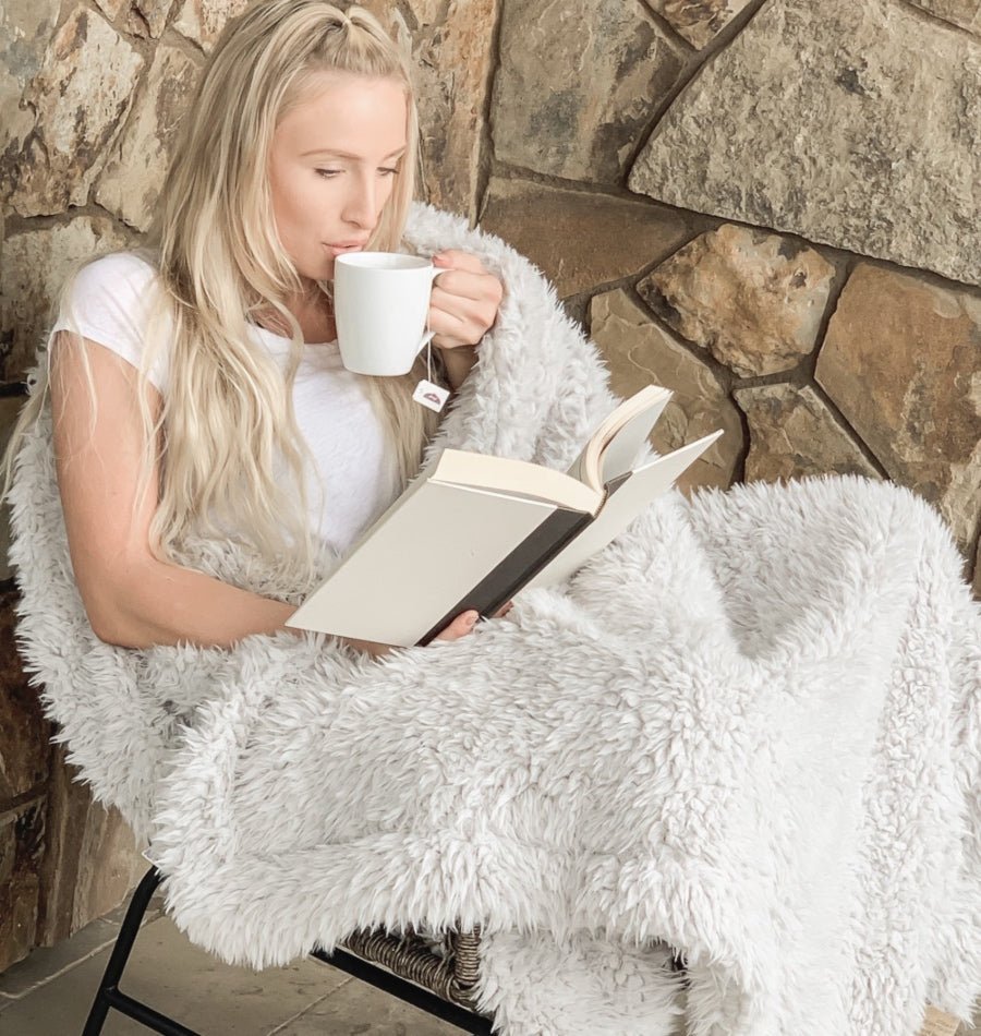 Woman reading a book and drinking from a mug wrapped in a fluffy white blanket against a stone wall.