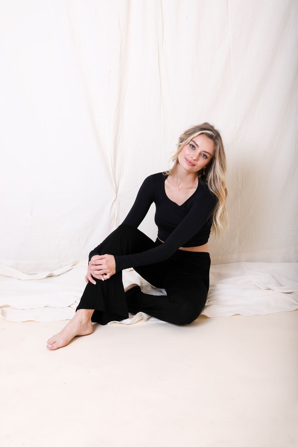 Woman in black outfit sitting on a white surface with a white curtain background