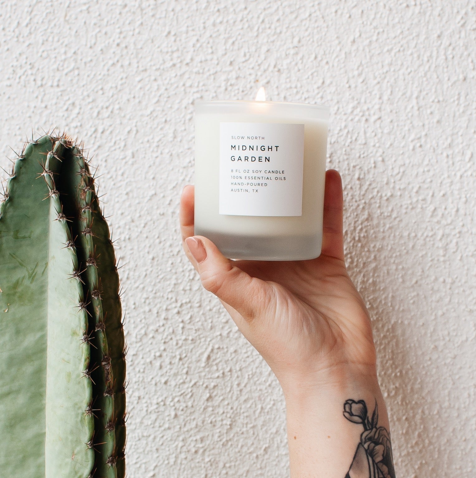 Hand holding a candle labeled 'Midnight Garden' next to a cactus against a light background